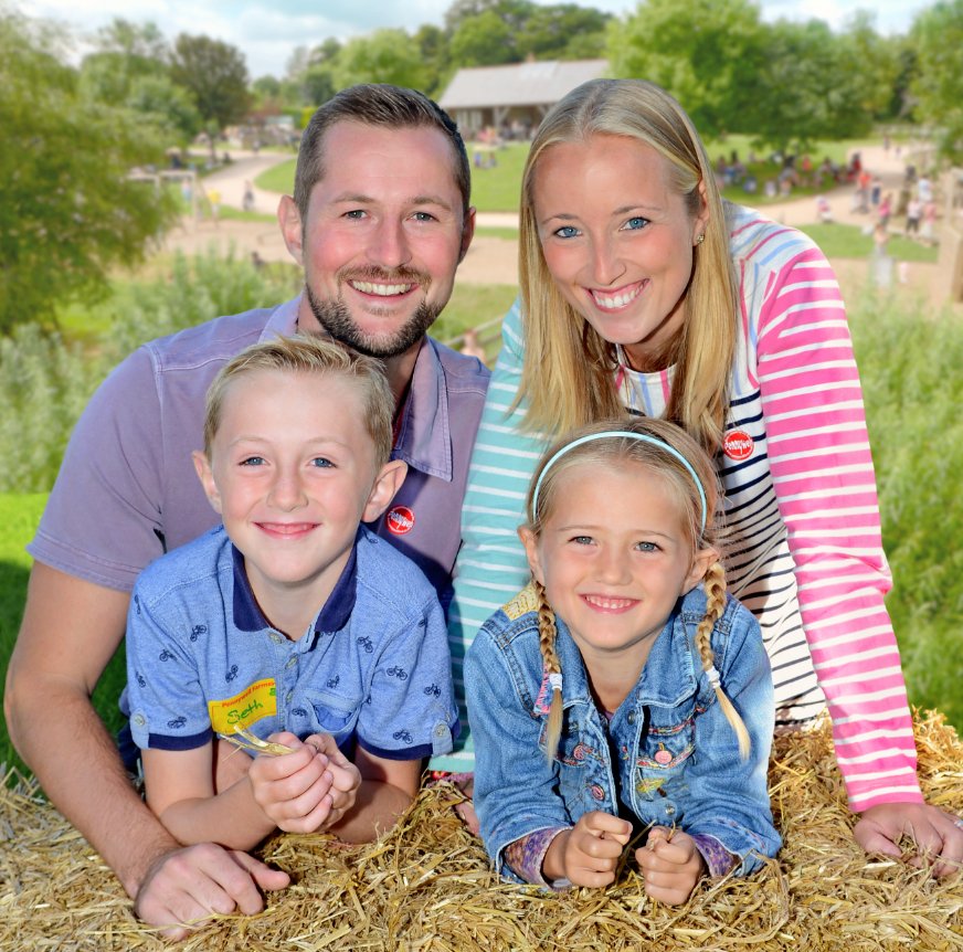 Family of four smiling outdoors, sitting on hay with green park background