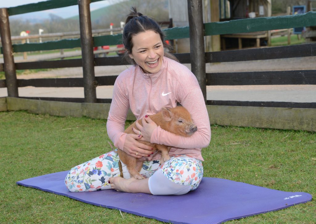 Woman in pink top and floral leggings sitting on purple yoga mat holding a small piglet outdoors