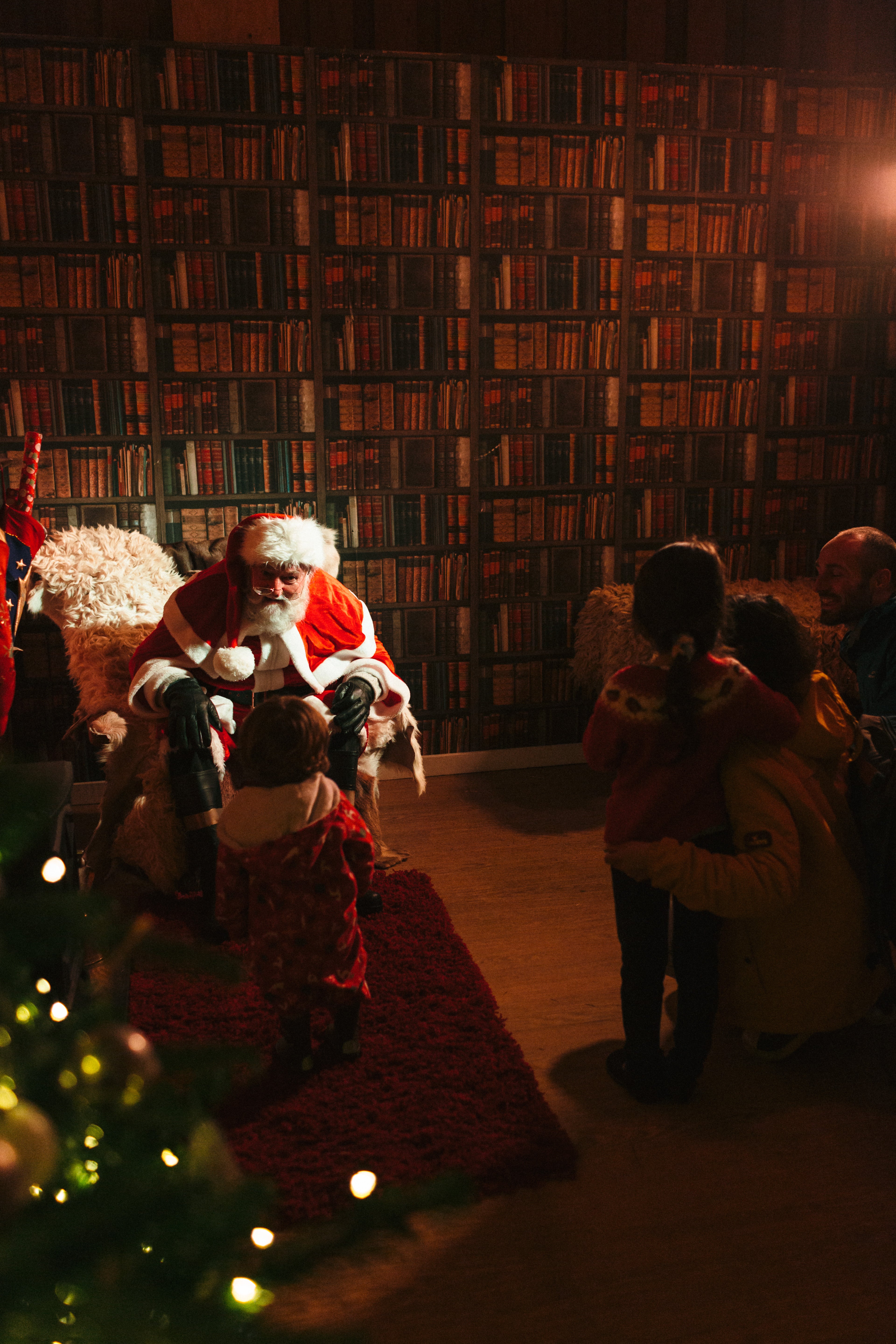 Santa Claus sits in a cosy setting, talking to children in festive clothing, with a bookshelf backdrop.
