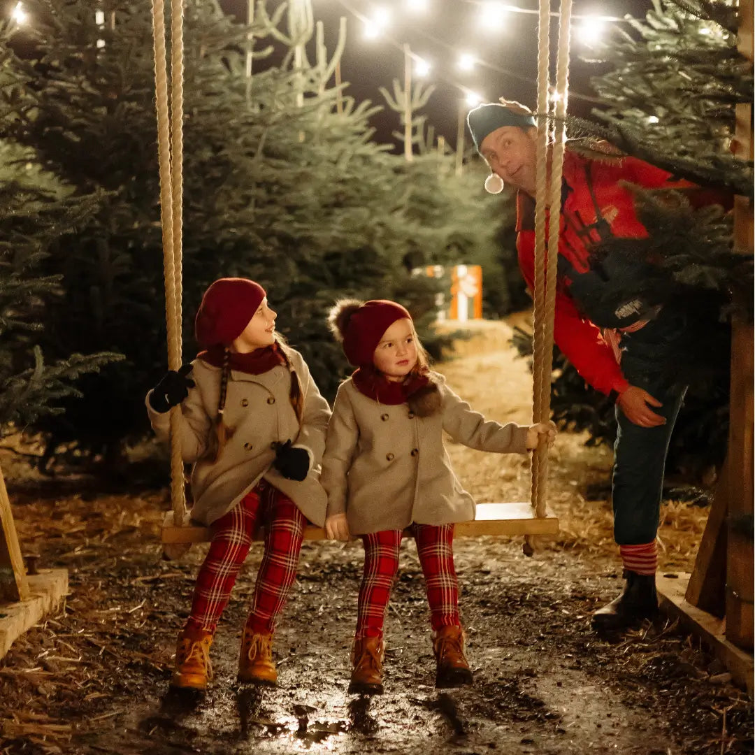 two girls in winter coats and hats sit on a swing, while a man in festive attire stands nearby among Christmas trees