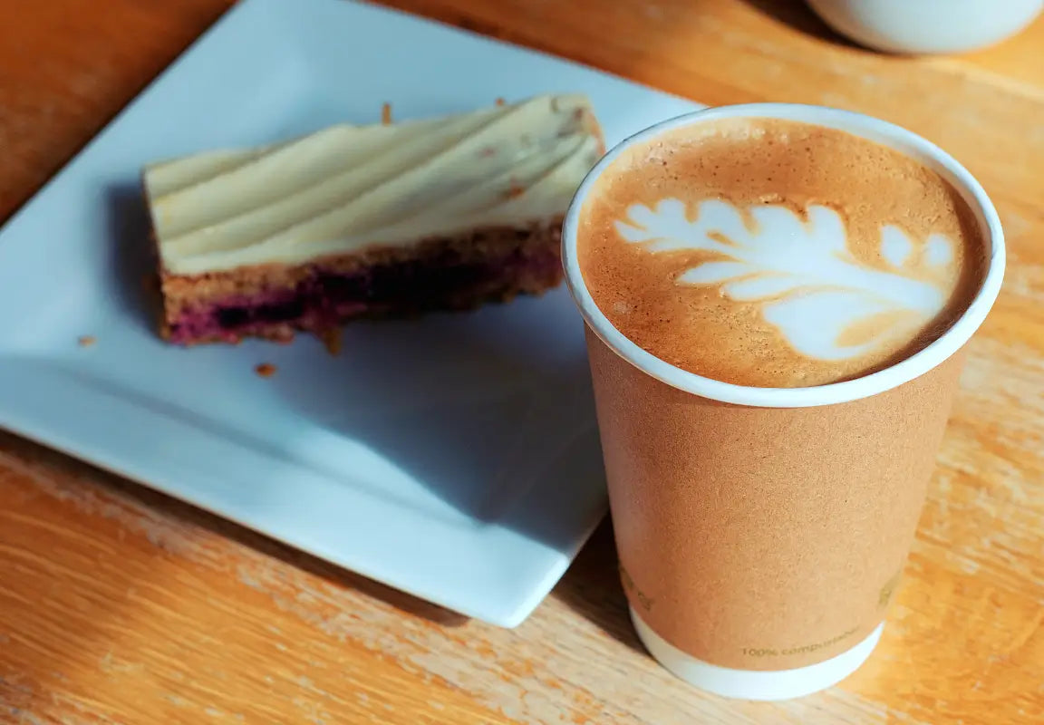 A cup of latte with leaf art beside a slice of cake on a white plate, set on a wooden table.
