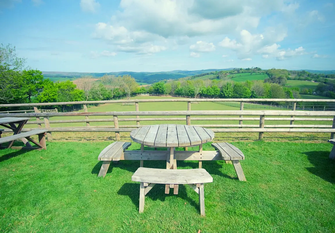 wooden picnic table and benches on grass, overlooking rolling hills and a wooden fence under a blue sky with clouds