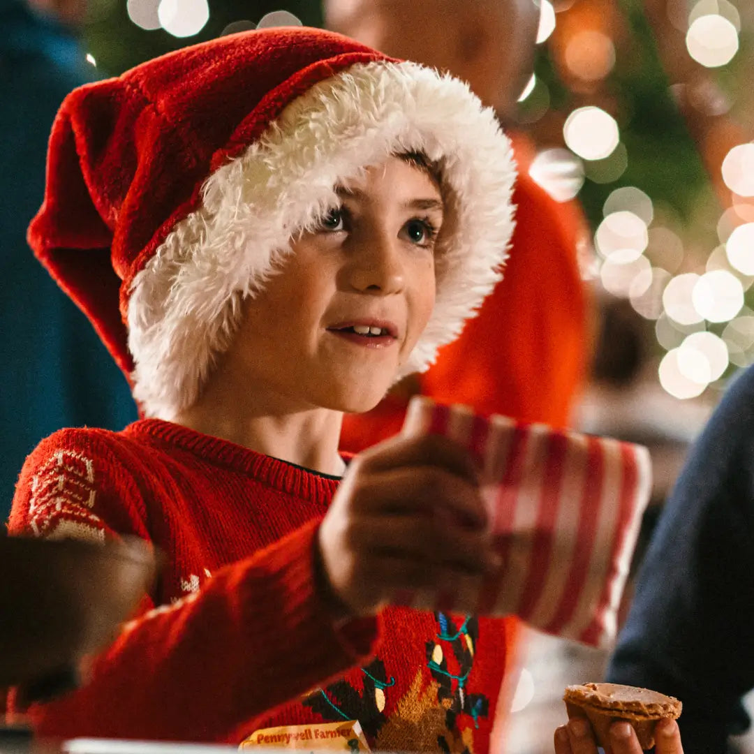 a young boy in a festive red sweater and Santa hat holds a striped gift while smiling, with Christmas lights in the background