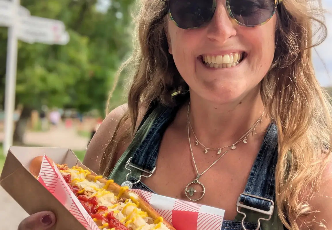 A woman in sunglasses smiles while holding a hot dog topped with mustard and ketchup in a park setting.