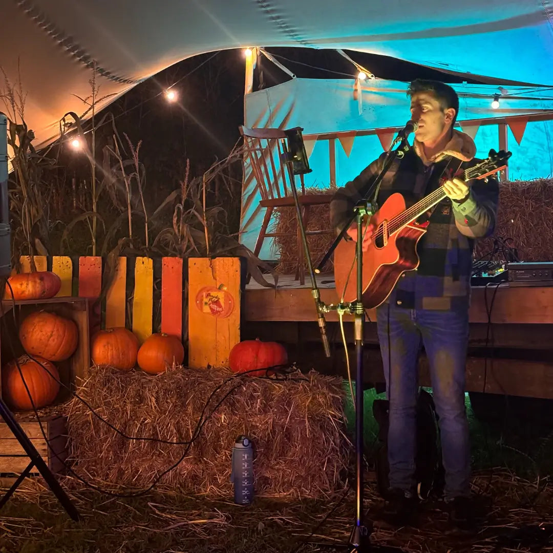 a musician performs with an acoustic guitar on a stage decorated with pumpkins and hay bales under string lights
