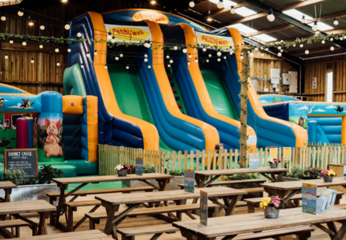 indoor play area featuring large inflatable slides and picnic tables, with decorative lights and a wooden fence