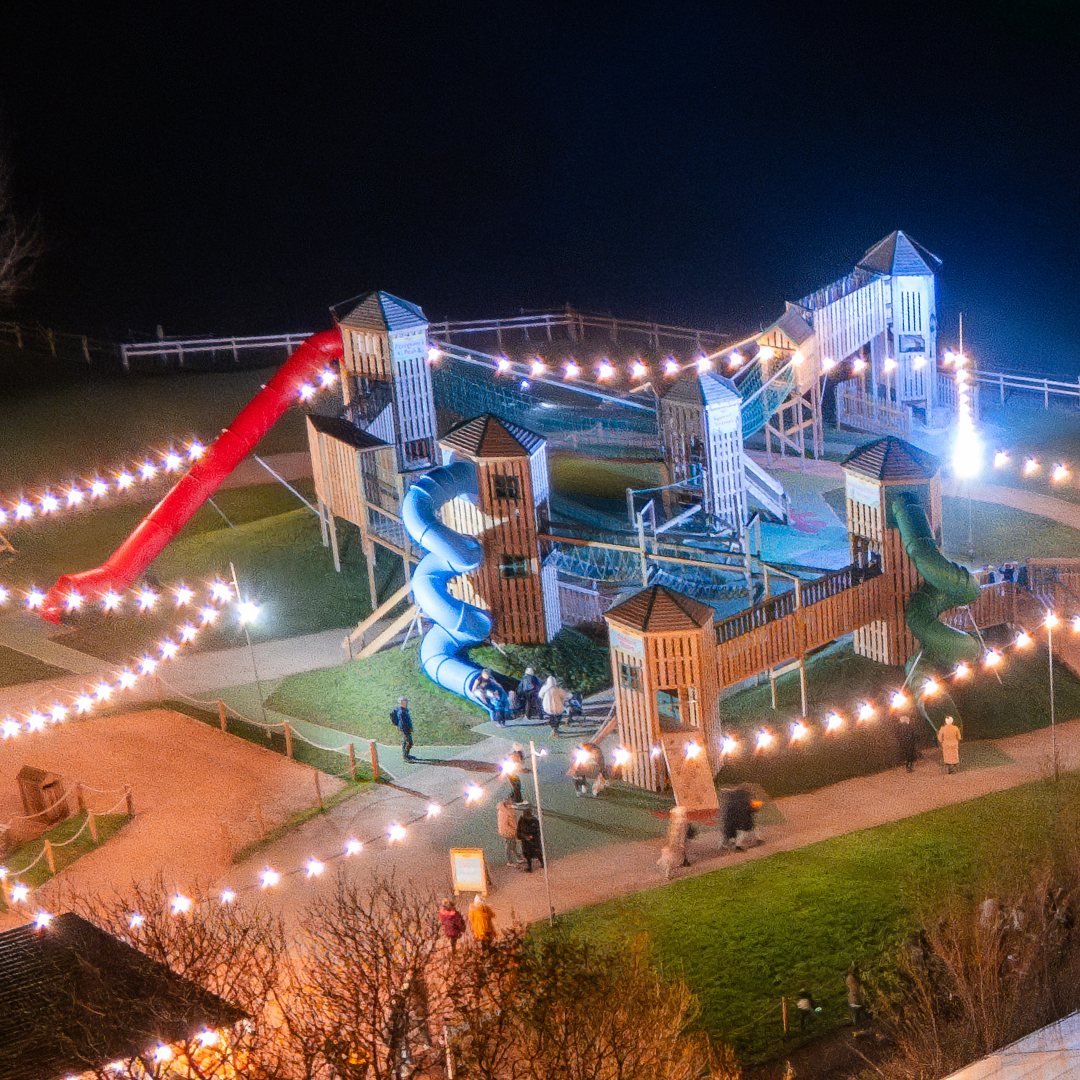 playground with red and blue slides, illuminated by string lights, with people walking around at night