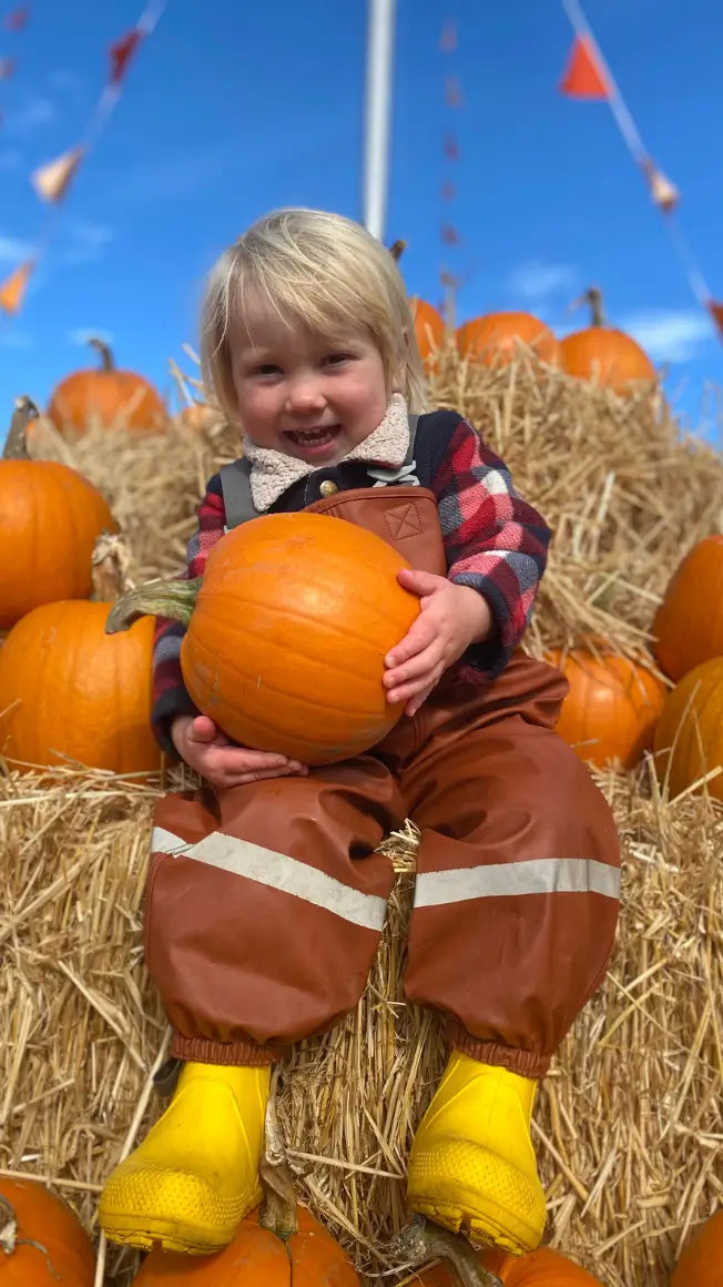 A young child in yellow wellies sits on hay bales, holding a pumpkin, with pumpkins in the background and a blue sky above.