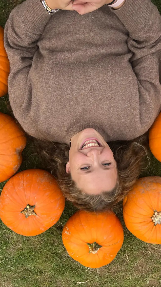 person lying on grass surrounded by pumpkins, smiling, wearing a brown sweater, with long curly hair