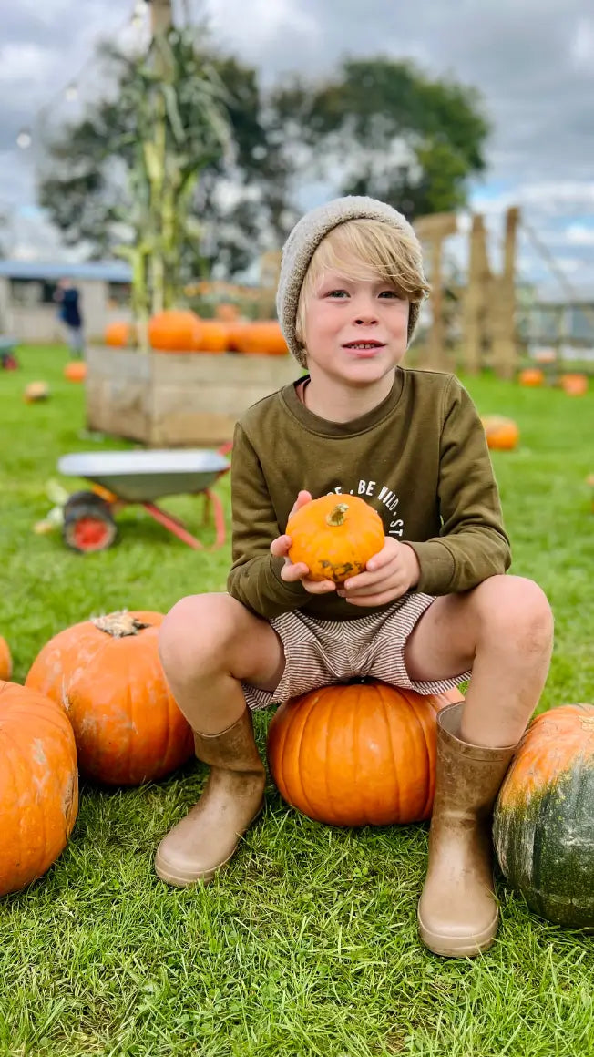 A boy sits on pumpkins, holding a small pumpkin, wearing a beanie and wellies, with a grassy field in the background.