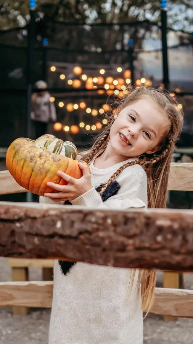 a young girl with braided hair smiles while holding a pumpkin, with fairy lights glowing in the background