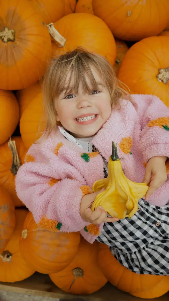 a smiling child in a pink jacket holds a yellow squash, surrounded by orange pumpkins in a farm setting