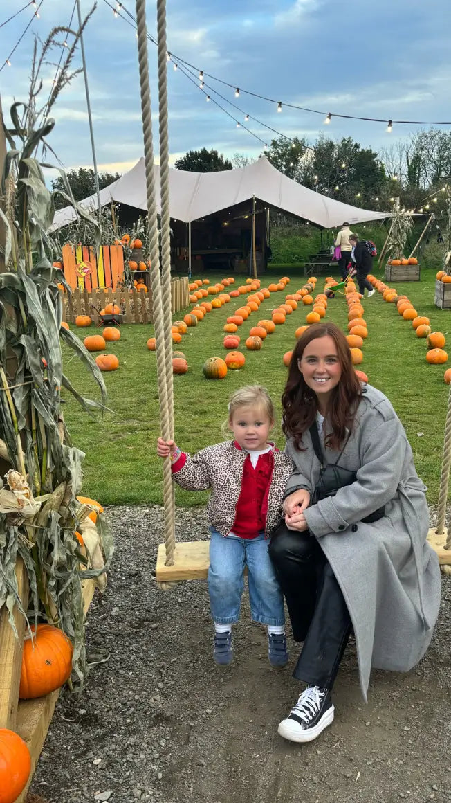 a woman and a child sit on a swing in a pumpkin patch, with pumpkins scattered across the grass and a tent in the background