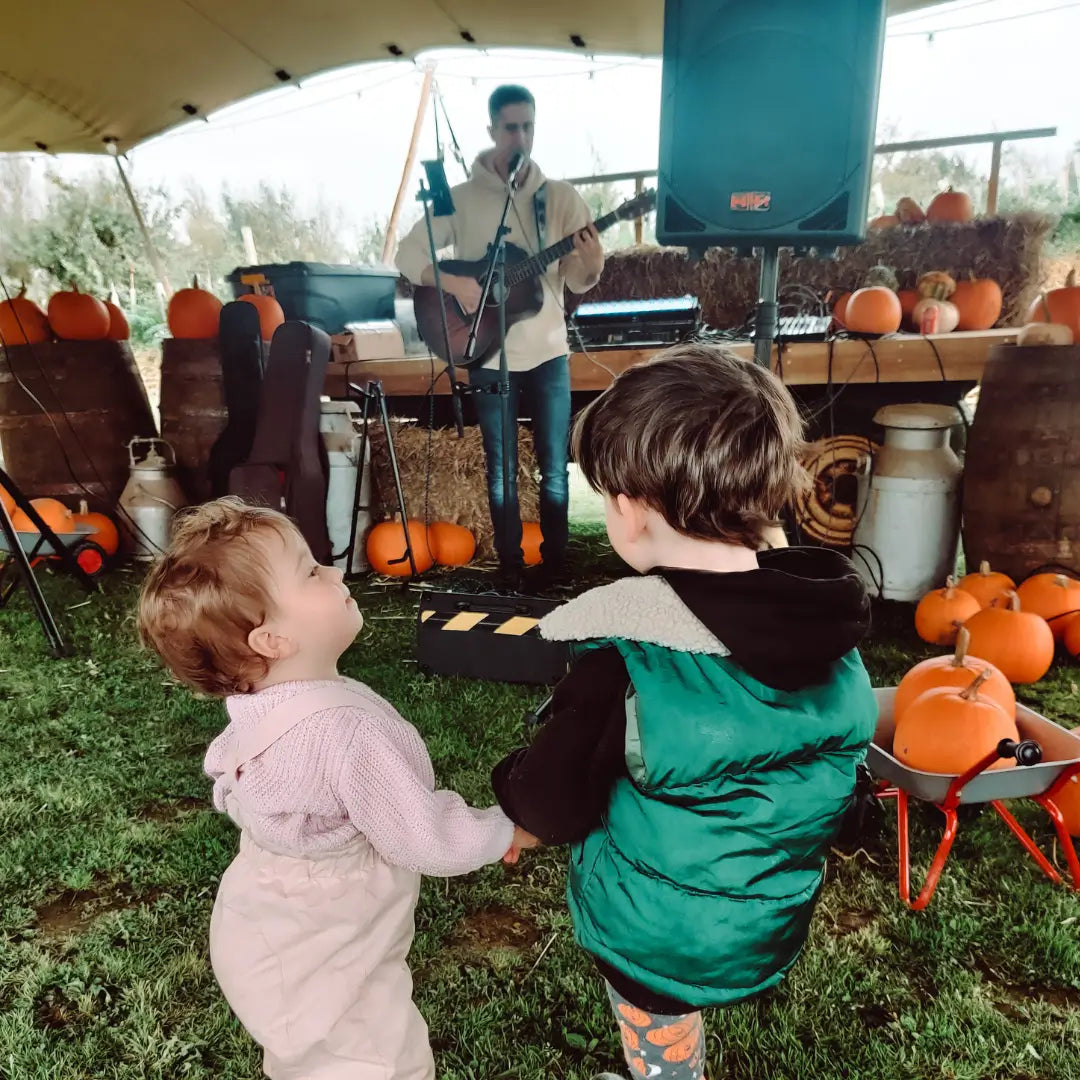 two children holding hands, watching a musician perform in a tent decorated with pumpkins and hay bales