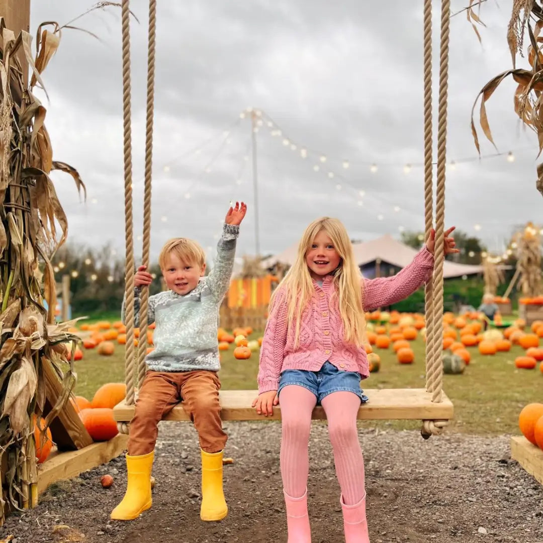 two children on a swing in a pumpkin patch, smiling and waving, with pumpkins and festive lights in the background