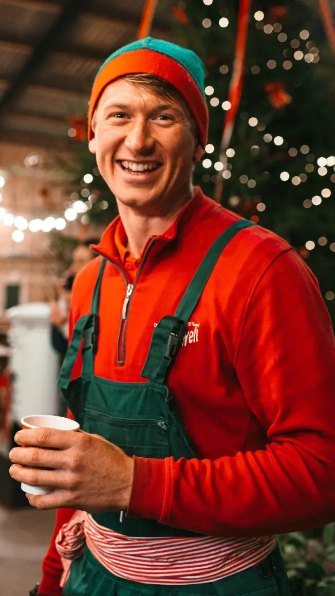 smiling man in red top and green overalls holding a cup, festive decorations in the background