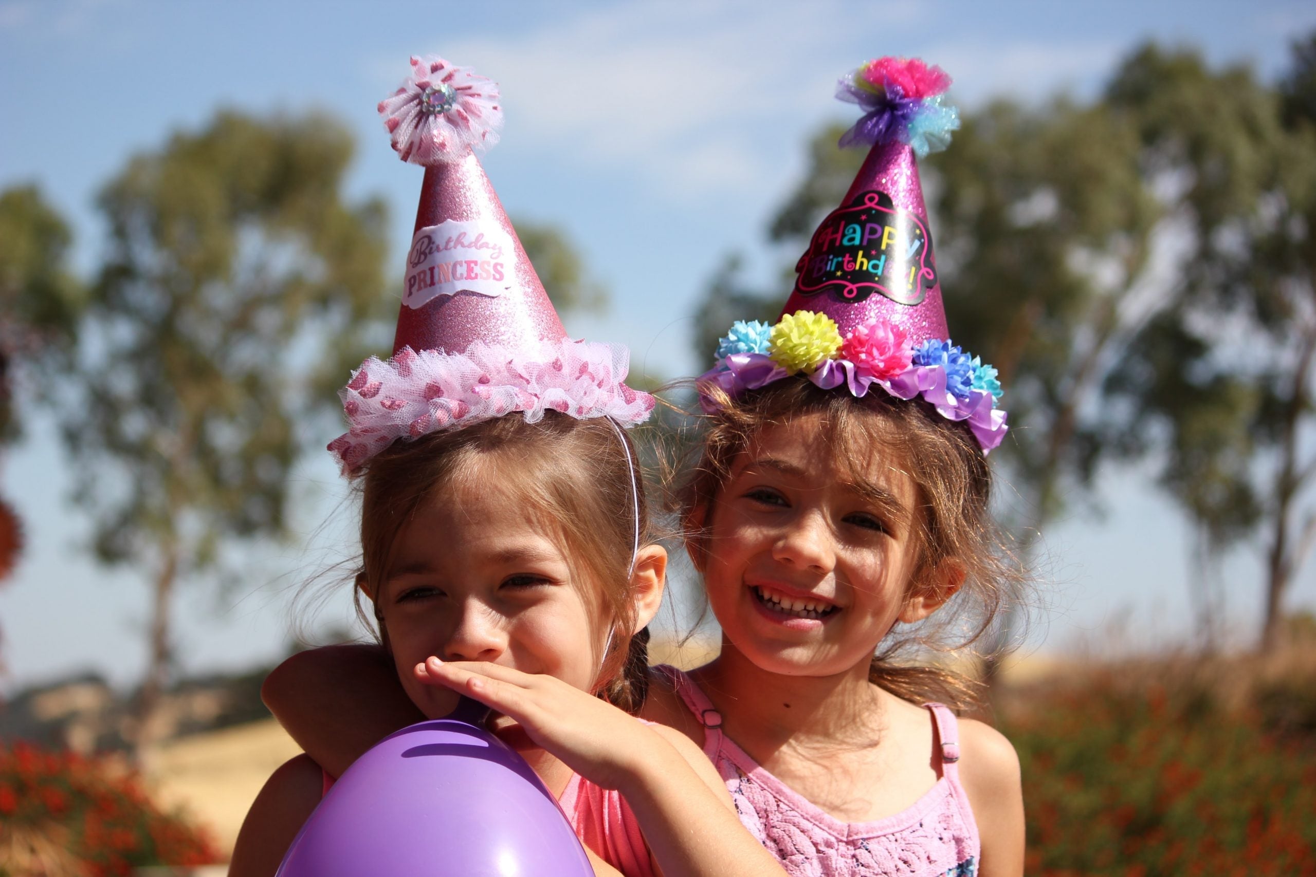 Two young girls wearing colourful birthday hats and smiling outdoors with a purple balloon