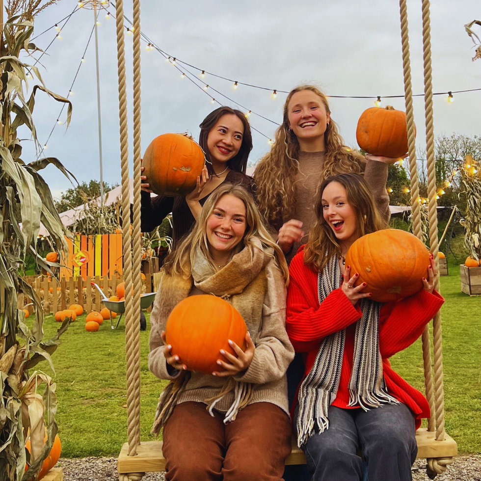 Four women hold pumpkins while sitting on a swing in a pumpkin patch, with festive lights and decorations in the background.
