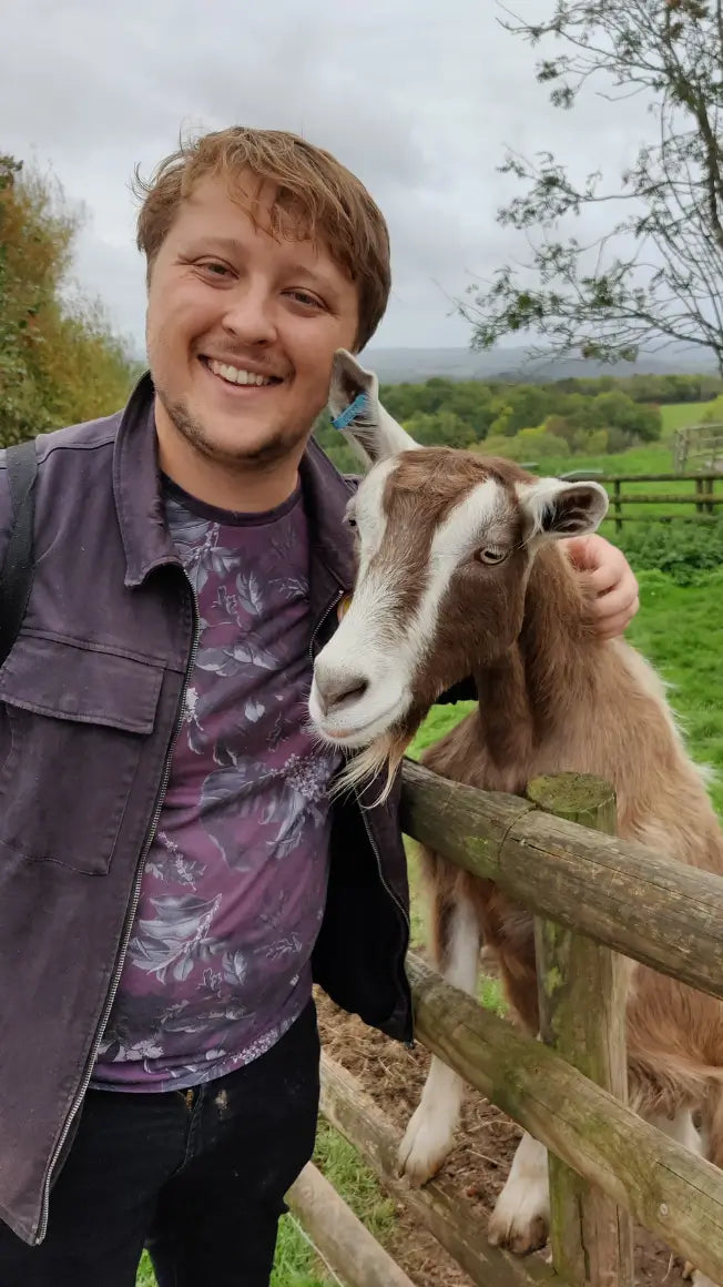 A smiling man stands next to a goat, both leaning against a wooden fence in a green field.