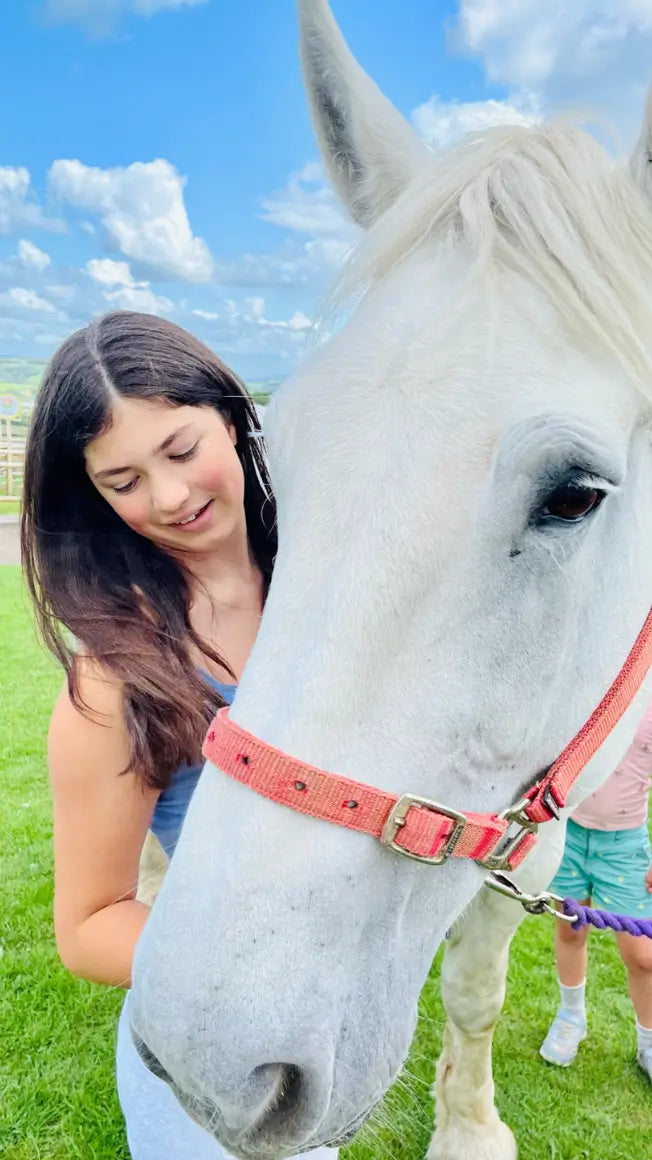 A young woman smiles while interacting with a large white horse on a sunny day in a grassy area.