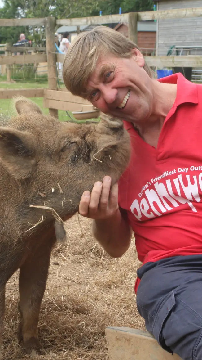 a man in a red shirt smiles while petting a small pig in a farm setting with straw on the ground
