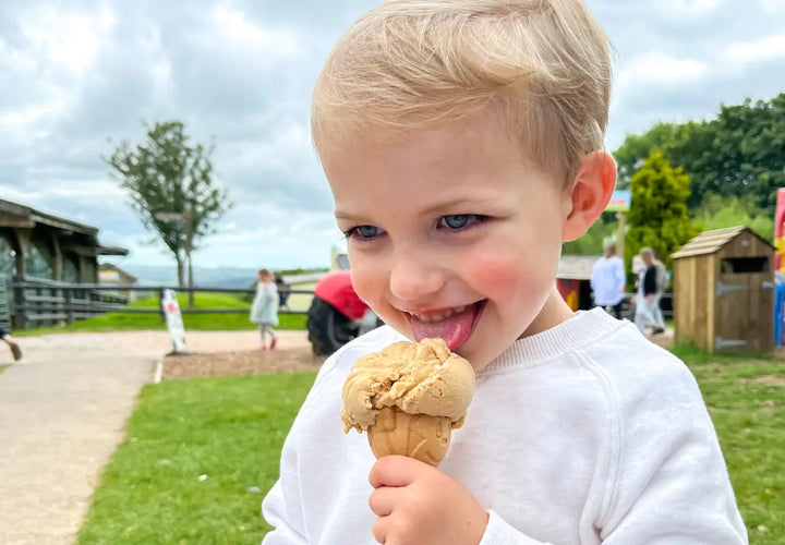 a young child with short blonde hair enjoys a cone of ice cream, smiling and licking the scoop in a park setting