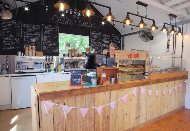 A café counter with a smiling barista, coffee machines, a menu board, and pastries displayed behind a glass case.