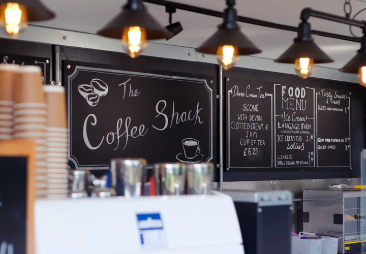 chalkboard menu at a coffee shop featuring drinks and snacks, with coffee cups and metal containers in the foreground