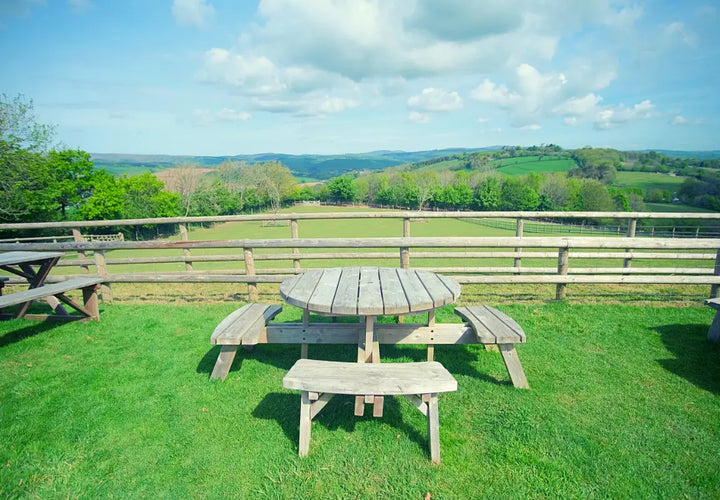 wooden picnic table and benches on grass, overlooking rolling hills and a wooden fence under a blue sky with clouds