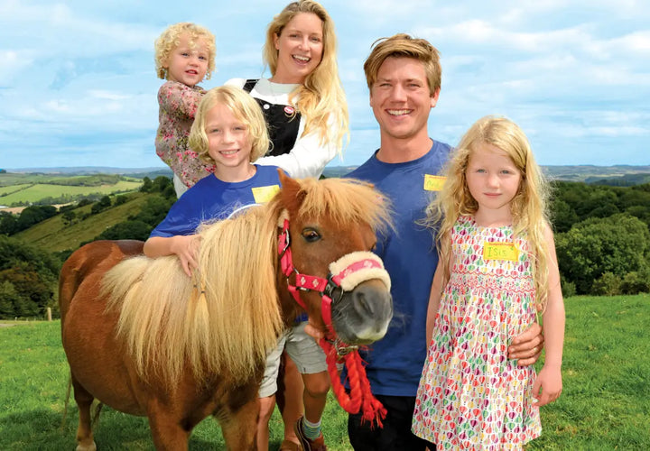 A family poses outdoors with a small pony, smiling against a backdrop of green hills and blue sky.