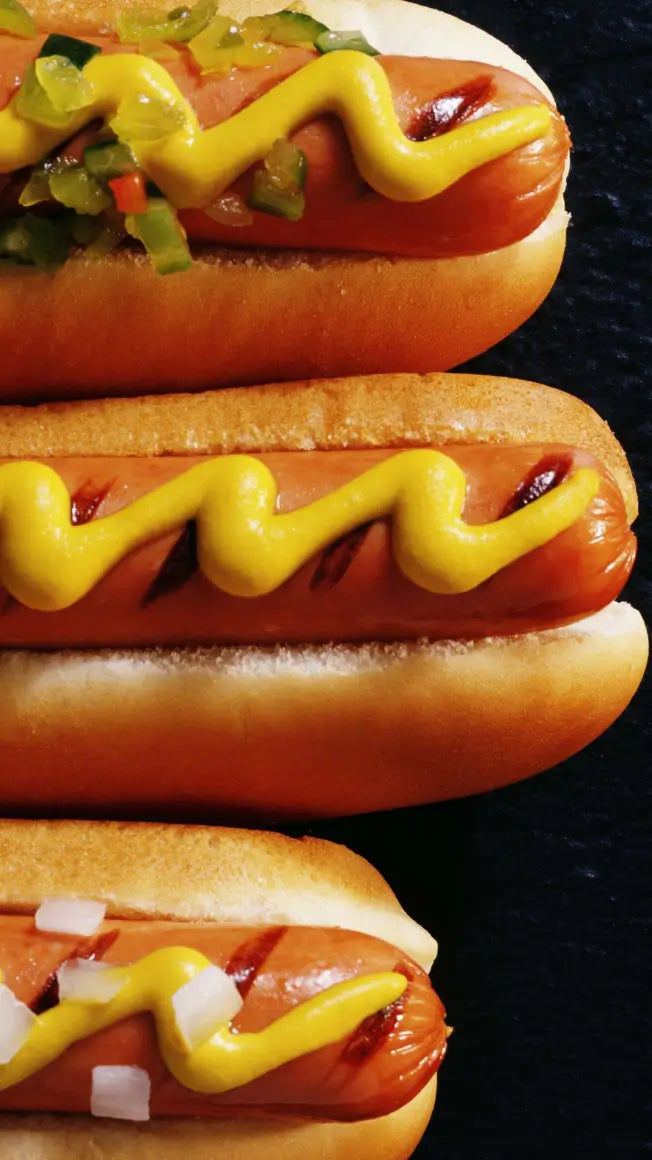 three hot dogs in buns, topped with mustard, diced onions, and chopped pickles on a dark background