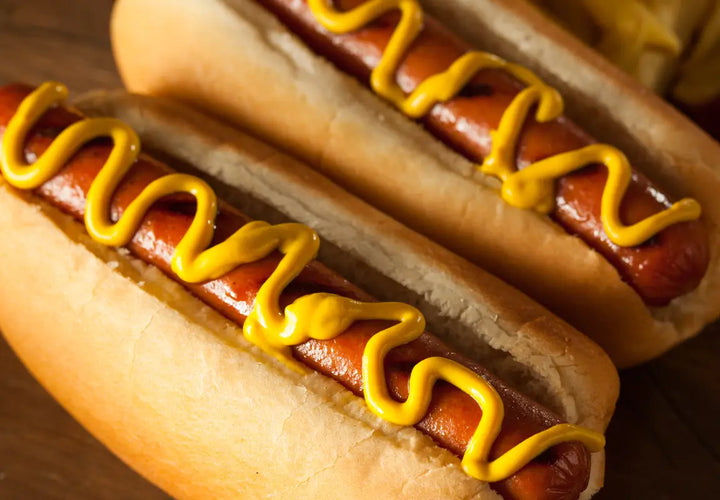 two hot dogs in soft buns topped with yellow mustard on a wooden surface