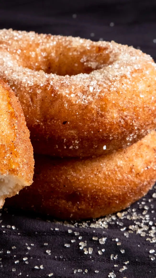 three sugar-coated doughnuts stacked on a dark surface, with granulated sugar scattered around them