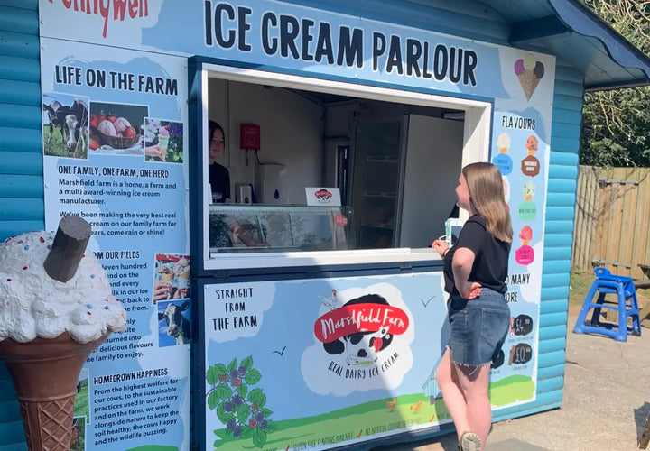 A woman stands at an ice cream parlour window, chatting with a staff member inside. Bright blue exterior with menu details.