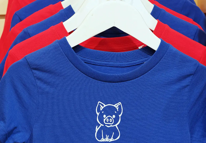 a close-up of blue and red t-shirts hanging on hangers, featuring a pig illustration on the blue shirt