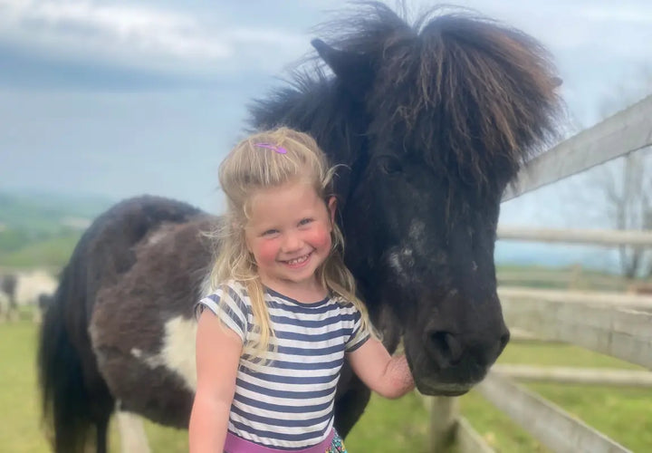 a young girl with blonde hair smiles beside a black and white pony in a field with a wooden fence in the background
