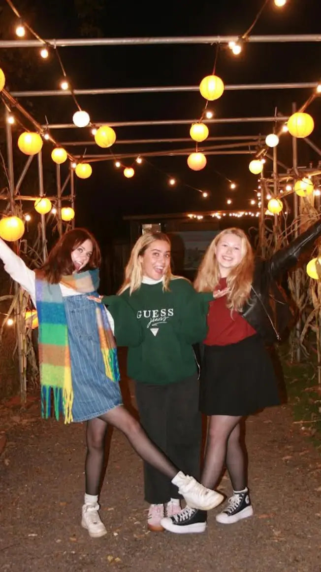 Three young women pose happily under string lights and lanterns, wearing casual outfits, in a festive outdoor setting.