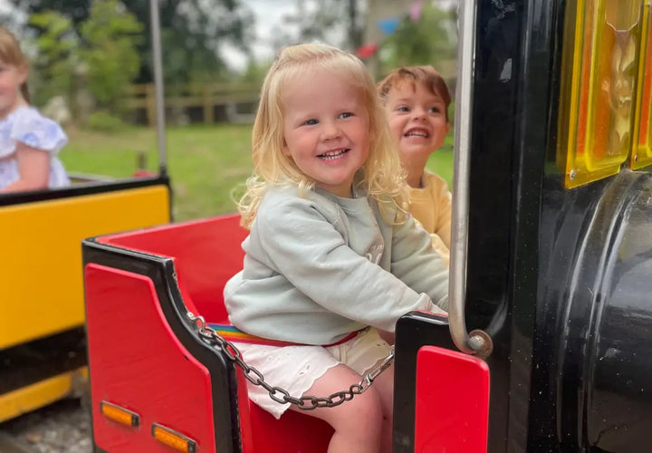 two children smiling on a small train ride, one girl with blonde hair in a grey jumper and a boy behind her