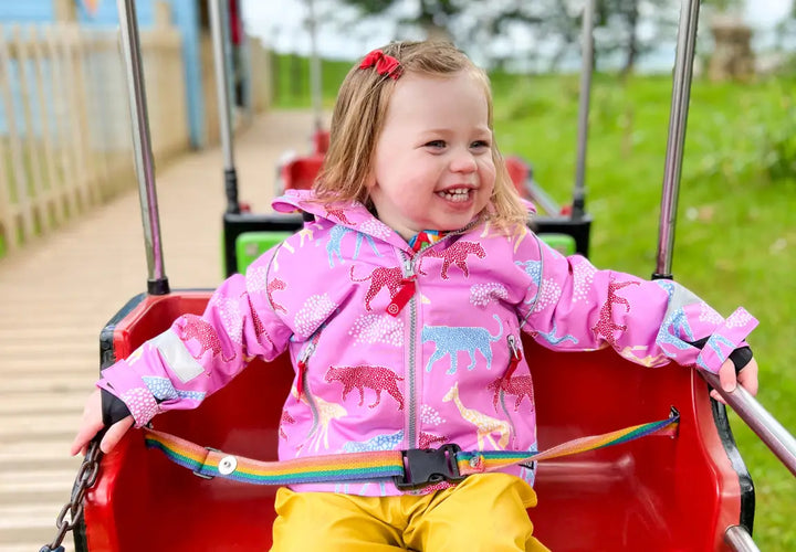 A smiling young girl in a pink animal-patterned jacket sits in a red ride, enjoying the outdoors.
