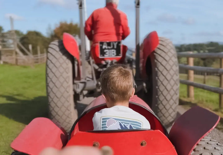 a child sits in a small red toy tractor, watching a larger red tractor driven by an adult in a field