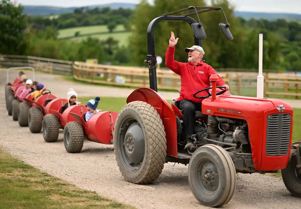 A man drives a red tractor, pulling a line of children in red carts along a gravel path in a rural setting.