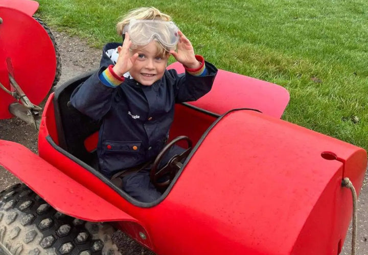 A young child wearing goggles smiles while sitting in a small red toy car on a grassy area.