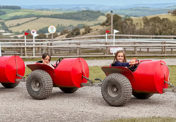 two children smiling in red pedal cars on a gravel track with green hills in the background