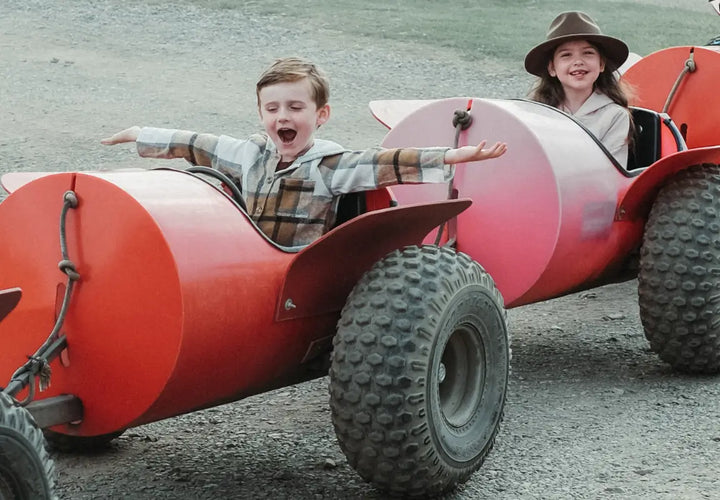 two children joyfully riding in toy cars, one boy with arms outstretched and a girl smiling beside him