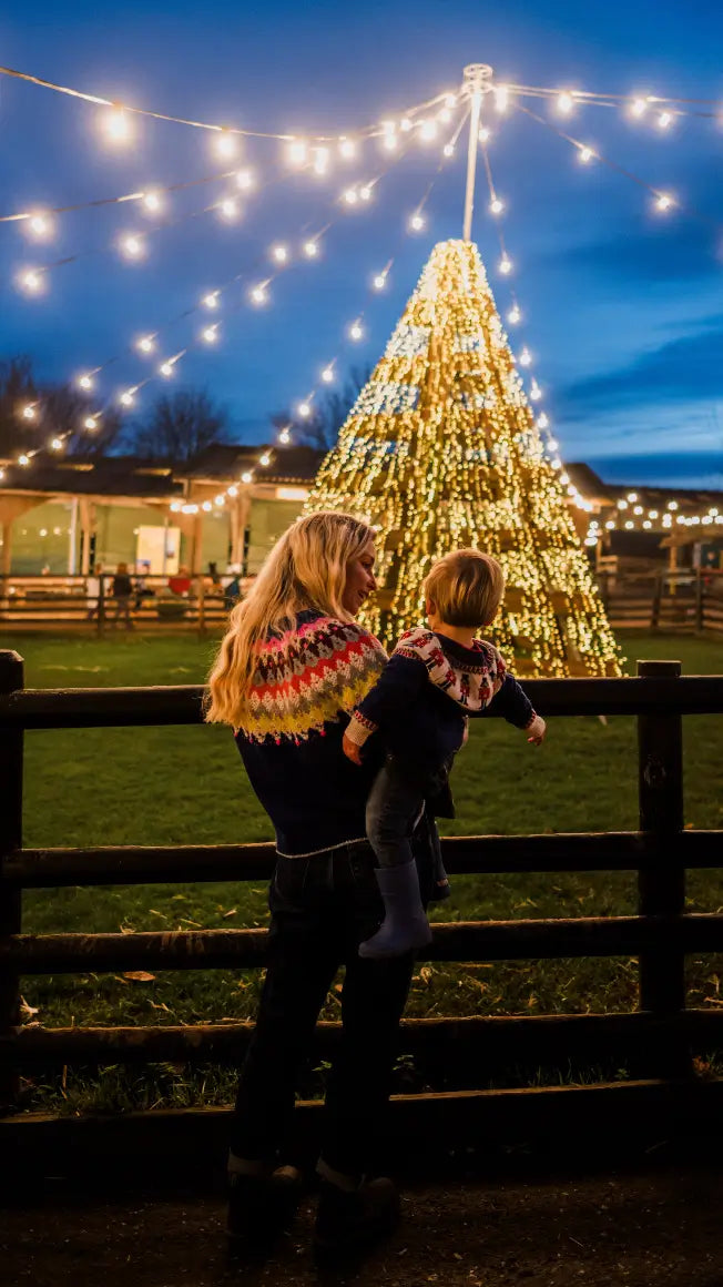 a woman holds a child, both in festive jumpers, in front of a lit Christmas tree at dusk with fairy lights around them