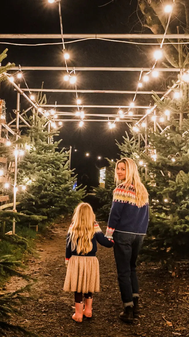 a woman and a girl walk hand in hand through a lit path lined with Christmas trees at night