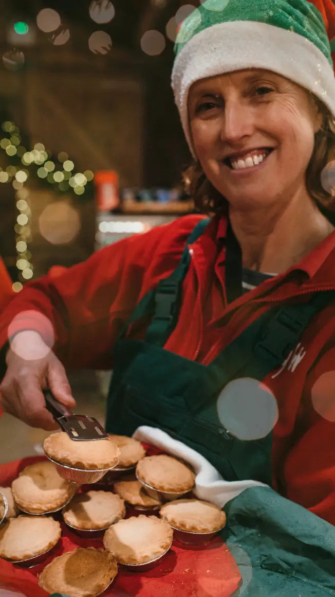 a woman in a festive hat holds a tray of mince pies, smiling in a warmly lit setting with holiday decorations