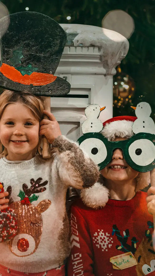 two children in festive jumpers holding props, smiling in front of a decorated Christmas backdrop