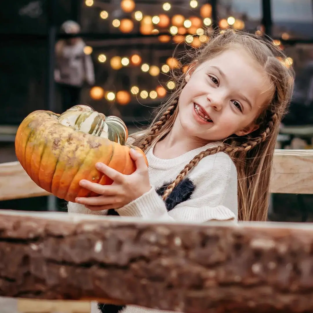 a young girl with braided hair smiles while holding a large pumpkin, with blurred lights in the background