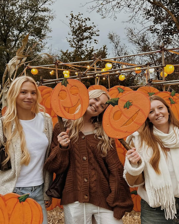 Three women hold pumpkin signs in front of a backdrop of smiling pumpkins, with autumn trees and lights in the background.